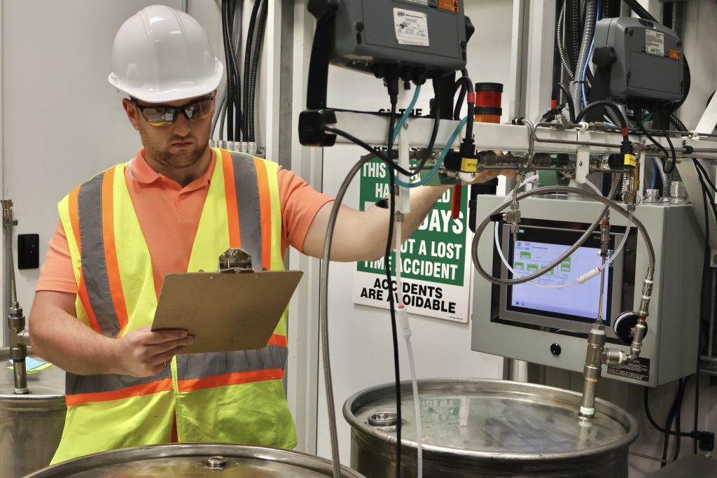 An Isowater employee wearing a hard hat and safety vest looks over manufacturing equipment with a clipboard in hand.